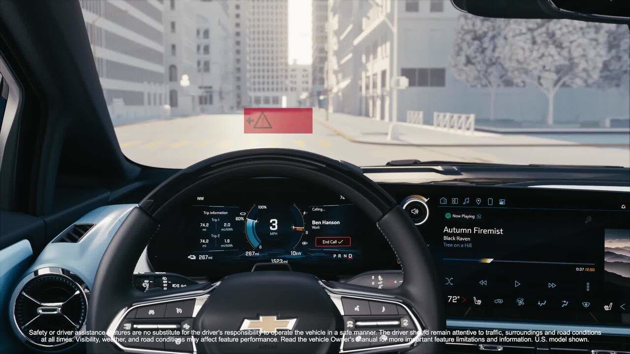 Vue intérieure depuis le siège du conducteur d'une Chevrolet Equinox EV. Un triangle d'avertissement rouge est affiché au-dessus du volant, indiquant un danger ou une alerte. Le tableau de bord numérique affiche diverses informations de conduite, et l'écran d'infodivertissement à droite affiche la lecture de musique. Une rue de ville est visible à travers le pare-brise.