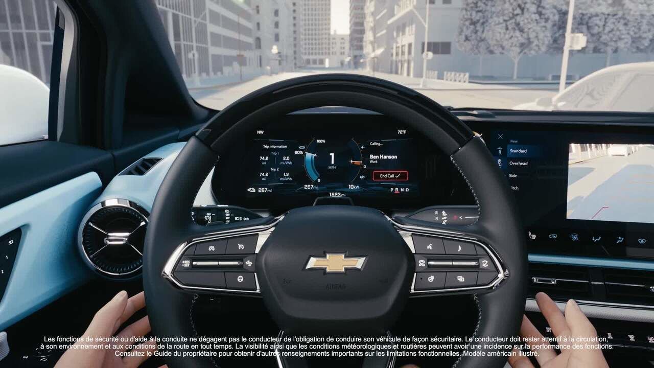 Les mains d'un conducteur sont sur le volant d'une Chevrolet Equinox EV, regardant à travers le pare-brise une rue de ville. Le tableau de bord numérique affiche des informations de conduite, y compris un indicateur de vitesse, la navigation et d'autres données du véhicule. Un texte en bas décrit les fonctions de sécurité et d'assistance en français et en anglais.