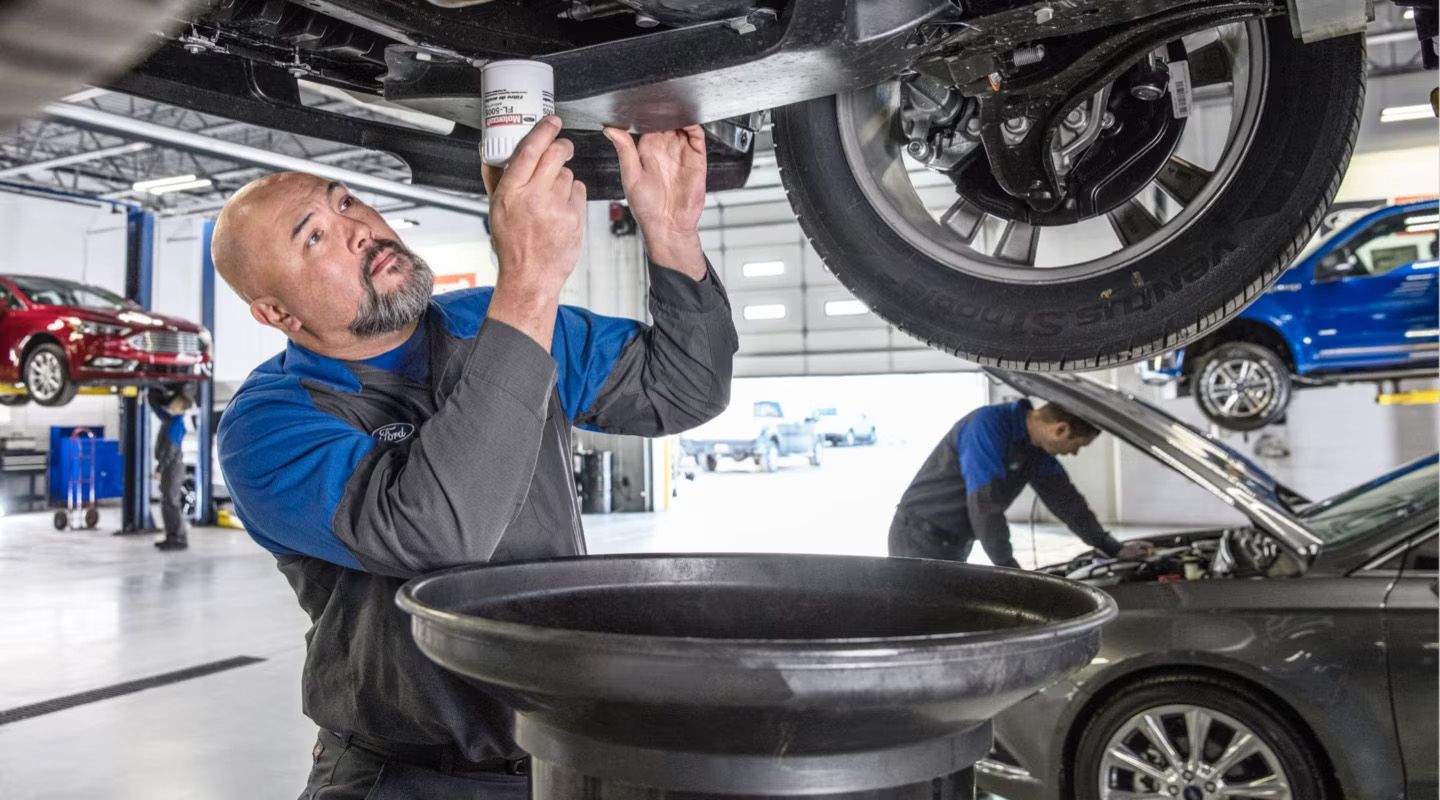 A mechanic, wearing a blue uniform, is working underneath a raised car in a service bay, holding what appears to be an oil filter. Another mechanic is visible in the background working on a different car.