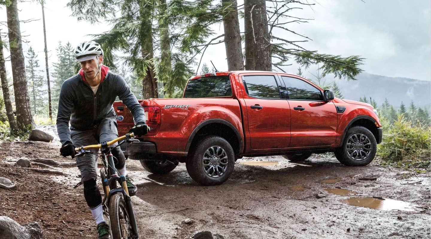 A man on a mountain bike rides on a muddy trail with a red Ford Ranger pickup truck parked behind him. Trees and mountains are in the background.
