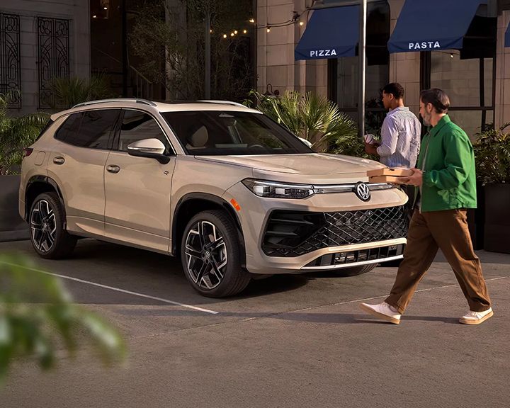 A beige SUV is parked in an urban setting at night. A man is walking towards the car, carrying a pizza box.