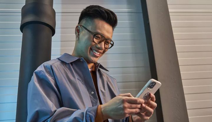 A smiling man wearing glasses looks at and uses his smartphone in an industrial or garage-like setting.
