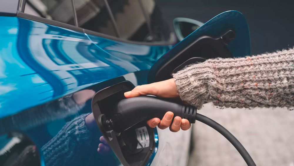 Close-up of a person's hand plugging a black charging cable into the side port of a teal electric car.