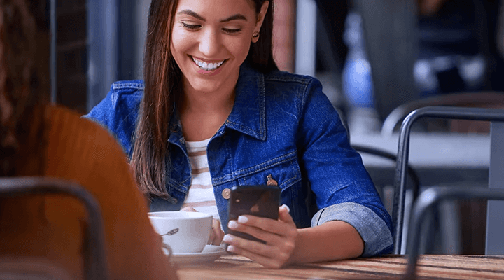 Une femme souriante assise à une table de café, regardant son téléphone intelligent.