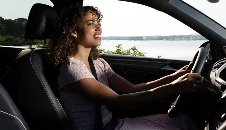 A young woman smiling while driving a car along a sunny coastal road with a view of the water.