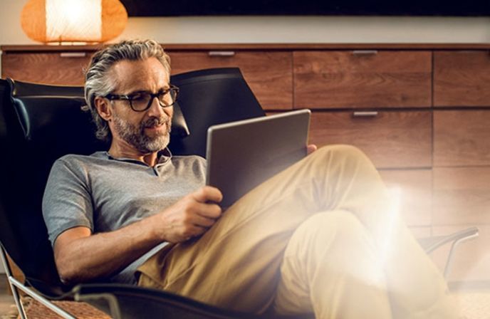 A mature man wearing glasses uses a digital tablet while sitting in a leather lounge chair.