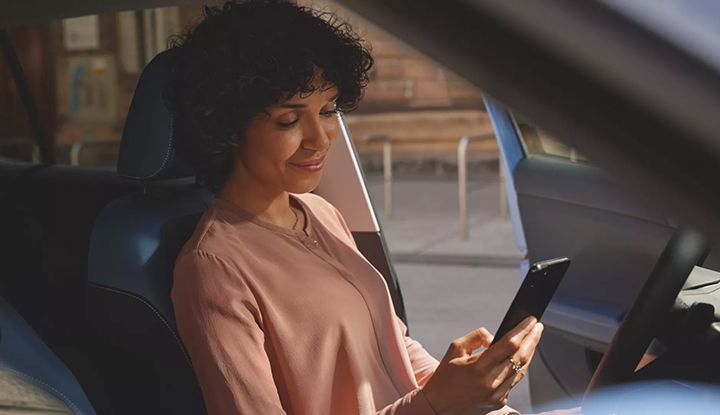 A smiling woman is sitting in the driver's seat of her car, looking at her smartphone.