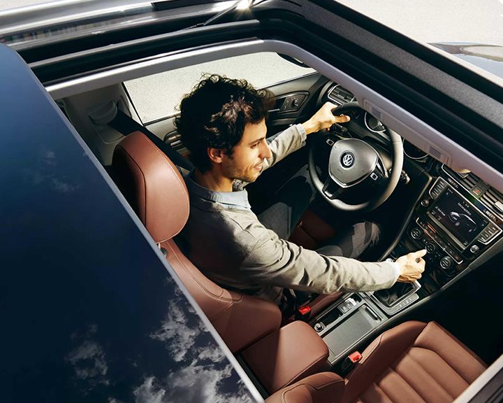 Vue plongeante d'un homme souriant conduisant une Volkswagen avec un toit ouvrant panoramique ouvert, montrant l'intérieur en cuir brun et le tableau de bord.