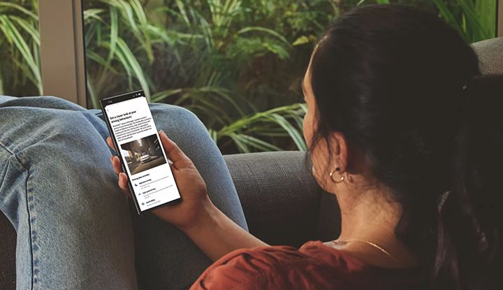A person is sitting comfortably on a couch, looking at a smartphone displaying DriveView data from the myVW app.