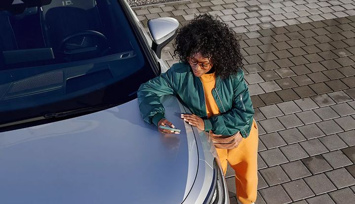 Overhead view of a woman using her phone and leaning on the hood of a silver Volkswagen ID. electric car.