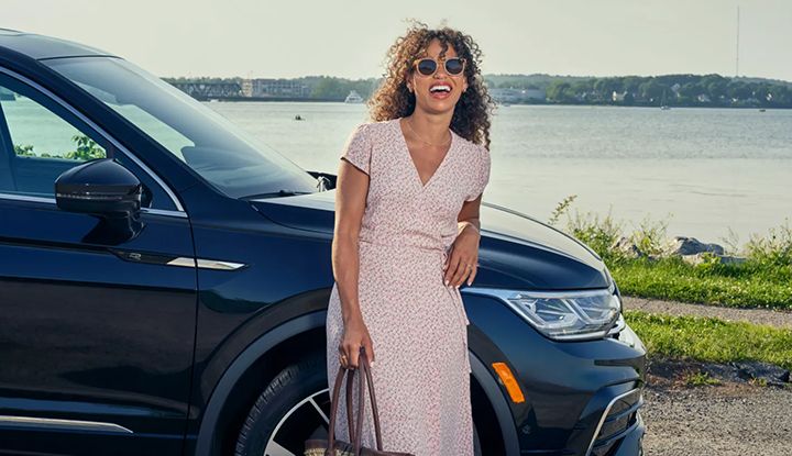 A smiling woman in a summer dress and sunglasses stands next to a dark SUV by a body of water.