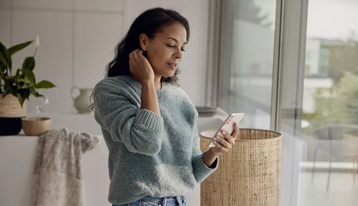 A woman standing near a window indoors is using a smartphone.