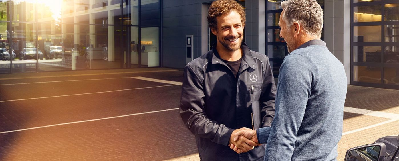 A smiling Mercedes-Benz technician shaking hands with a mature customer outdoors.