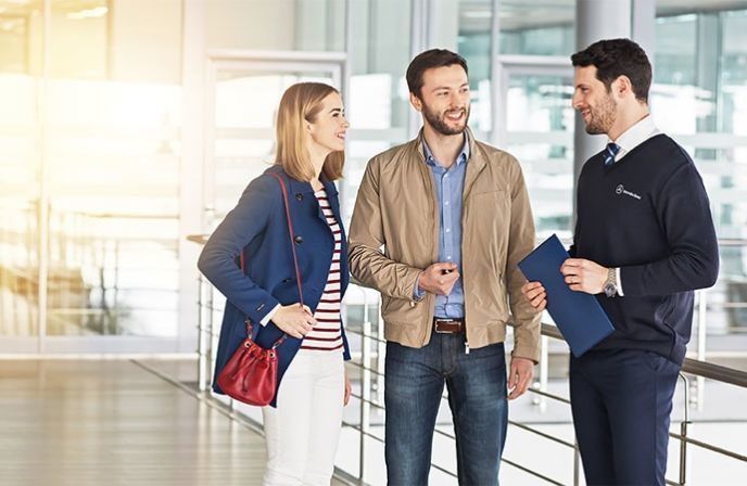 A smiling couple talking to a Mercedes-Benz advisor holding a blue folder.