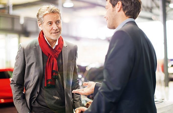 A mature man wearing a red scarf talking to an advisor inside a car dealership.