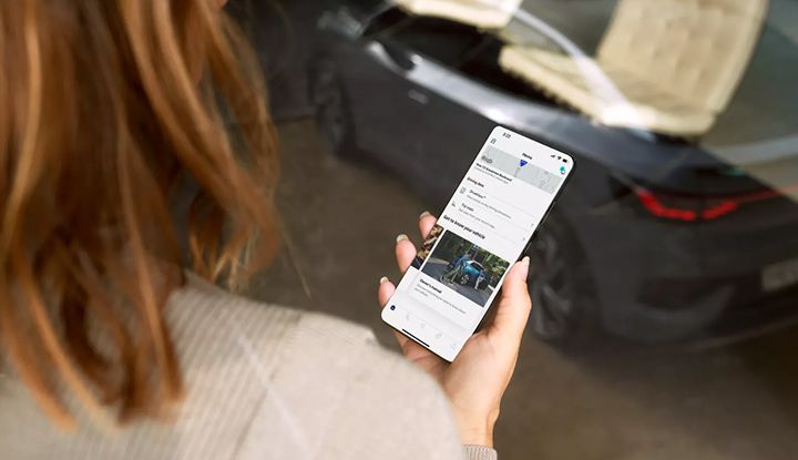 A person is holding a smartphone that displays the myVW app's Digital Owner's Manual, with a car visible in the background.