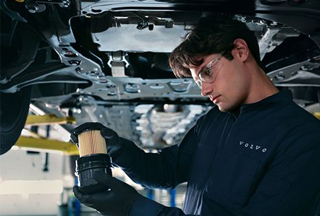 A technician in a Volvo uniform and safety goggles holds an oil filter beneath a car lifted on a hoist in a garage.