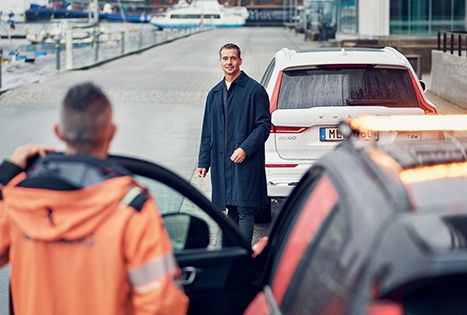 A man in a dark coat smiles at a tow truck operator in an orange uniform (viewed from behind) next to his broken-down car near a dock.