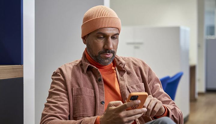 A bearded man wearing an orange beanie and a turtleneck is looking at and using a smartphone.