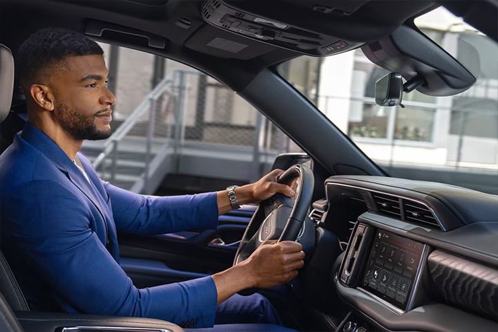 A man in a dark blue suit driving a vehicle, looking ahead, with the infotainment screen displaying vehicle service applications.