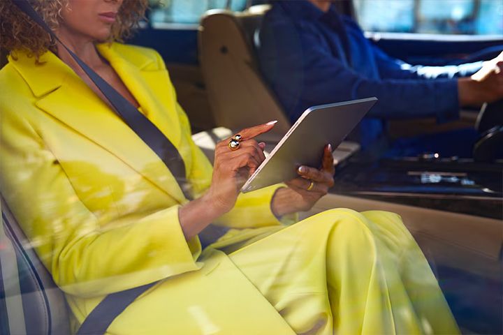 A woman in a bright yellow suit seated in the passenger seat of a car, using a tablet or small laptop.