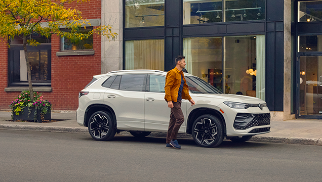 A man in a mustard yellow jacket walks past a modern white SUV parked along a city street in front of a brick and glass building.