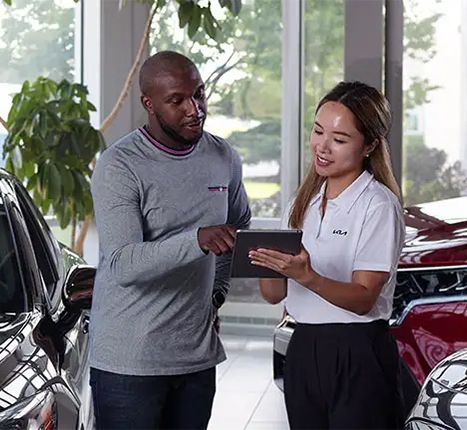 A car salesperson is showing a customer information on a tablet in a car dealership showroom.