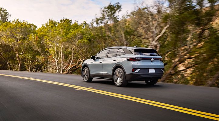 Rear view of a light blue Volkswagen ID.4 electric SUV driving on a winding country road surrounded by trees.