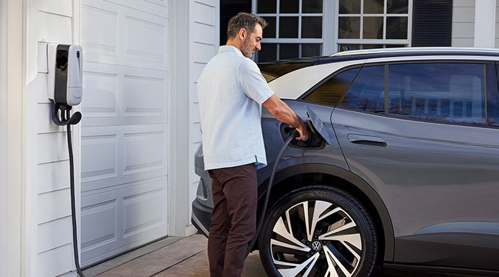 A man is charging a gray Volkswagen ID.4 electric SUV with a home charging station next to a garage.