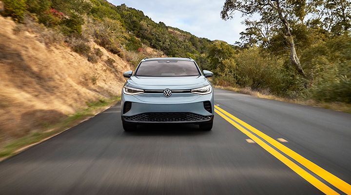 Front view of a light blue Volkswagen ID.4 electric SUV driving on a mountain road with double yellow lines.