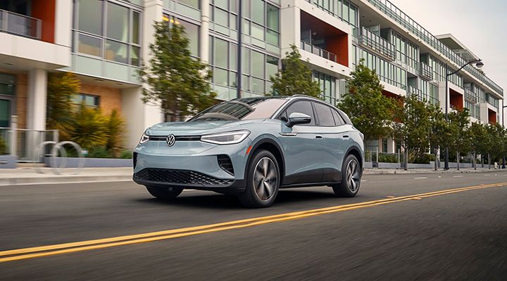 Un VUS électrique Volkswagen ID.4 bleu clair roule dans une rue de ville bordée de bâtiments modernes.