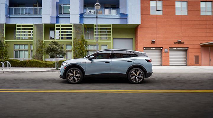 Side view of a light blue Volkswagen ID.4 electric SUV driving on an urban street lined with colorful buildings.