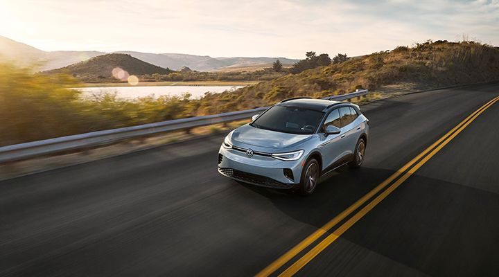 A light blue Volkswagen ID.4 electric SUV driving on a sunny coastal highway next to a lake or body of water.