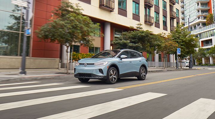 A light blue Volkswagen ID.4 electric SUV drives on a city street, passing a crosswalk.