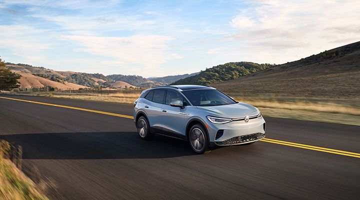 A light blue Volkswagen ID.4 electric SUV driving on an open country road with grassy hills in the background.