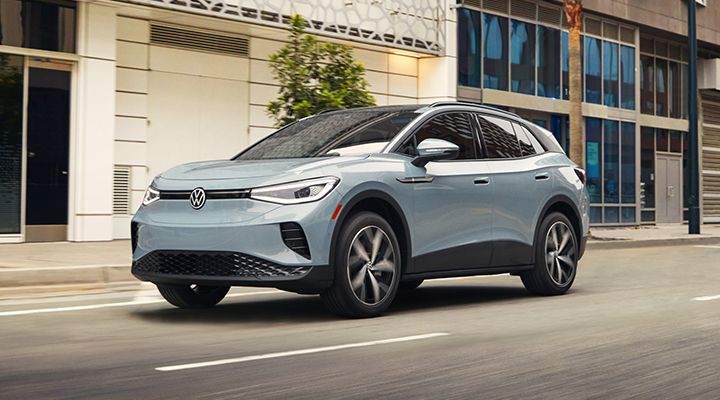 A light blue Volkswagen ID.4 electric SUV drives on a city street lined with modern buildings.