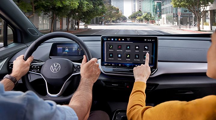 Interior view of a Volkswagen ID.4 showing hands on the steering wheel and a passenger interacting with the large central touchscreen.