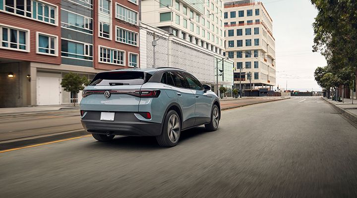 Rear view of a light blue Volkswagen ID.4 electric SUV driving on a city street lined with modern buildings.