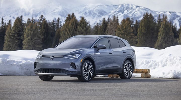 A gray Volkswagen ID.4 electric SUV is parked on a snowy road with mountains and pine trees in the background.