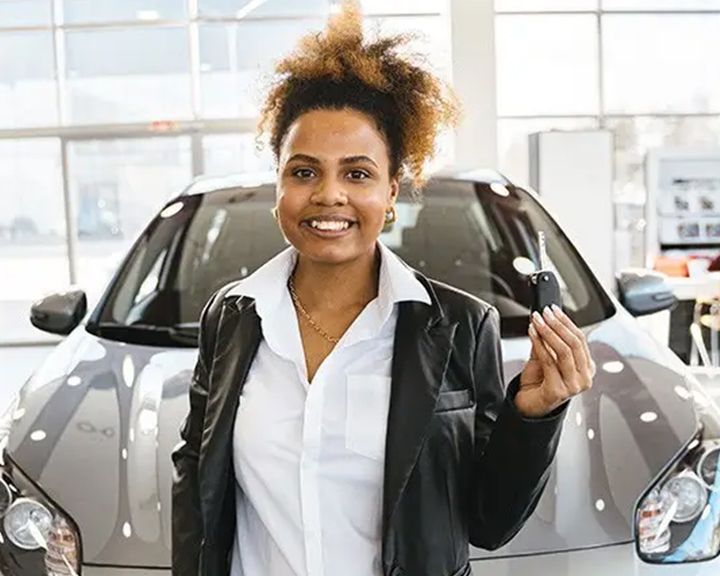 A smiling woman holds up the keys to her new car, standing in front of the vehicle in a dealership showroom.