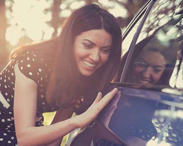 A smiling woman touches the fender of a car, examining the paint.