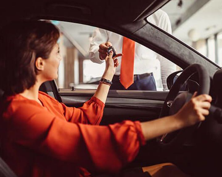 A salesperson hands car keys to a customer sitting in the driver's seat of a new vehicle.
