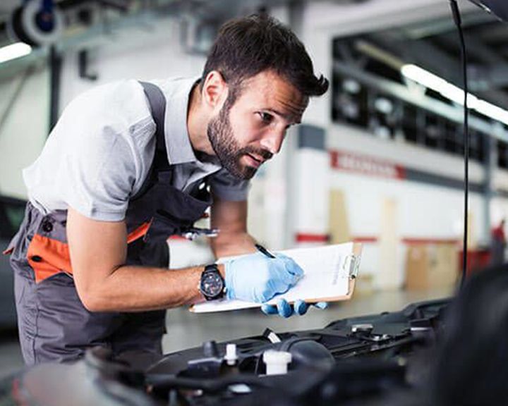 A mechanic inspects a car engine, taking notes on a clipboard.