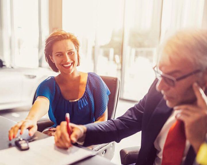 A smiling woman signs vehicle purchase documents while sitting across from a male salesperson.