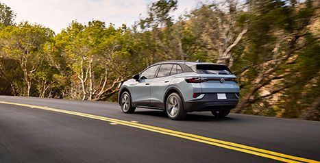 Rear view of a light blue Volkswagen ID.4 electric SUV driving on a winding, tree-lined country road.