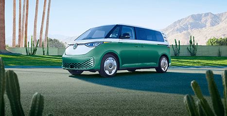A two-tone green and white Volkswagen ID. Buzz electric van is parked on a road lined with palm trees and cacti.
