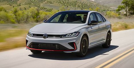 A white Volkswagen Jetta GLI sedan with red accents and a black roof drives on a country road under a sunny sky.