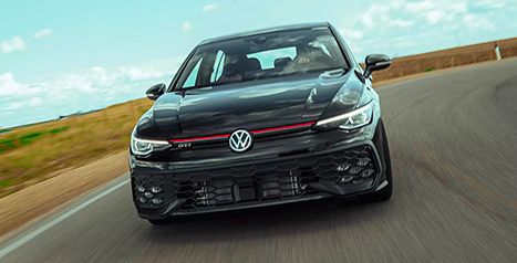 Black Volkswagen Golf GTI, front view in motion, driving on a country road under a blue sky.