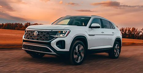 White Volkswagen Atlas or Atlas Cross Sport, front three-quarter view in motion, driving on a dirt road at sunset.