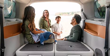 A family is sitting in the open rear cargo area of a van on a beach, smiling and talking.